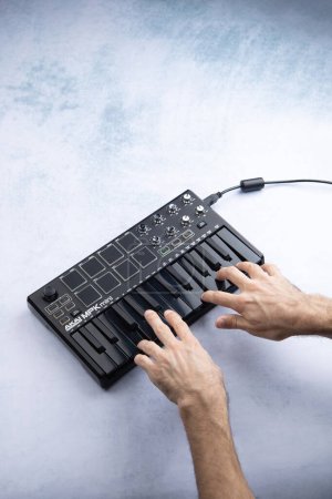 Téléchargez les photos : Une main d'homme joue les touches d'un mini clavier midi noir Akai mpk mk2, sur fond blanc et bleu - en image libre de droit