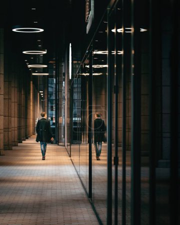 Straßenbild eines Mannes im langen Mantel, der nachts in London im langen Korridor mit Spiegelung im Glas weggeht