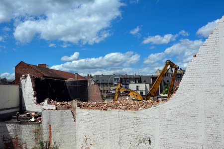 Photo for Behind the dilapidated brick wall of the building, excavators are demolishing the house against the background of a bright sky - Royalty Free Image