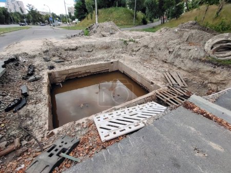 Téléchargez les photos : Un trou carré a été creusé sur la route avec des tuyaux d'égout inondés d'eau. Travaux de réparation de revêtements et d'assainissement de mauvaise qualité - en image libre de droit