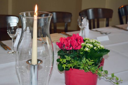 A pot with pink cyclamen flowers on a table near a glass vase with a burning candle. A row of chairs in the back blurred background
