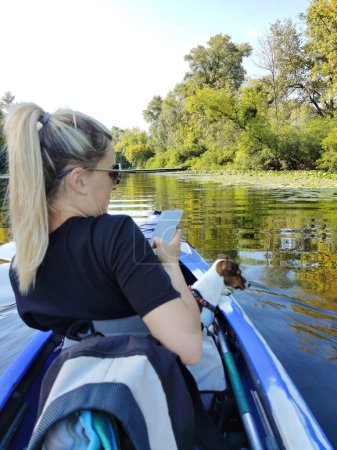 Eine junge Frau segelt mit ihrem Jack Russell Terrier Hund in einem Boot auf dem Fluss und macht ein Foto von ihr auf ihrem Handy