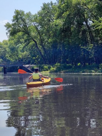 A teenage boy is kayaking on a river against a background of trees and a small bridge. Rear view. Active life and rest