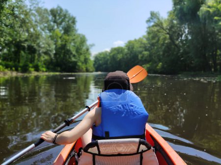 A teenage boy is swimming in a life jacket in a kayak on the river against the background of trees. Rear view. Active life and rest