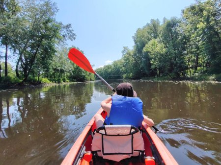 A teenage boy is swimming in a life jacket in a kayak on the river against the background of trees. Rear view. Active life and rest