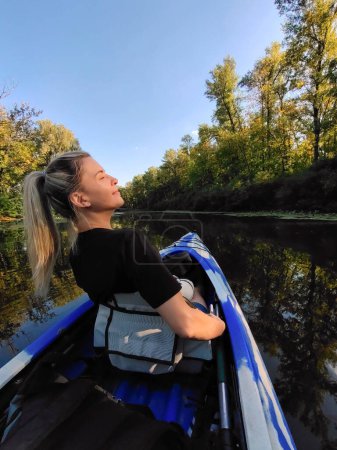 Eine junge blonde Frau schwimmt in einem Boot und blickt lächelnd vor dem Hintergrund des Flusses und der Bäume zurück. Tourismus, Sport und Erholung im Freien
