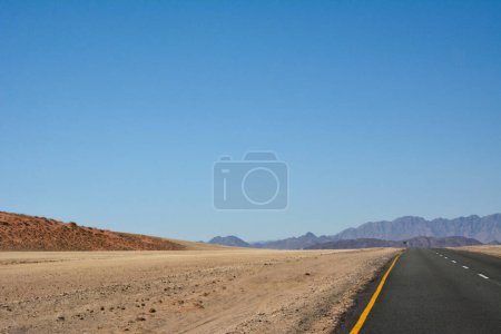 Camino de asfalto en perspectiva en el desierto bajo cielo azul claro. El camino conduce a montañas y colinas distantes