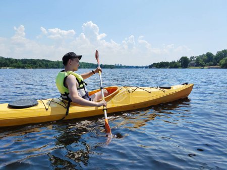 A young guy is swimming in a kayak with a paddle in a life jacket on a wide river. Active sports in the summer in nature