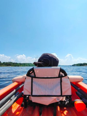 Ein kleiner Teenager schwimmt entspannt in Schwimmweste und Mütze auf einem Boot auf dem Fluss. Rückansicht