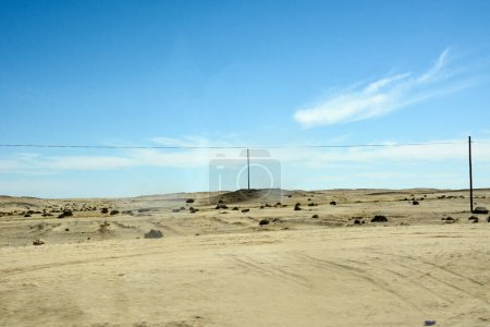 Un paysage chaud dans un désert sec. Les poteaux de lignes électriques sont au premier plan. En arrière-plan se trouvent des montagnes lointaines et un ciel bleu clair