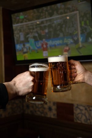 Foto de Dos hombres brindando con vasos de cerveza frente a un televisor con un televisor mostrando un partido de fútbol en la pantalla. - Imagen libre de derechos