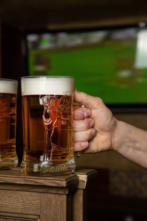 Foto de Dos hombres brindando con vasos de cerveza frente a un televisor con un televisor mostrando un partido de fútbol en la pantalla. - Imagen libre de derechos