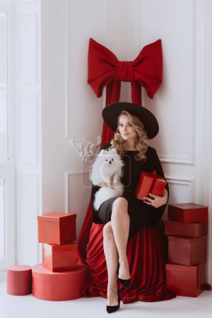 Foto de Hermosa mujer elegante en vestido festivo negro y sombrero con perro blanco en las manos y regalos de Navidad. Enfoque selectivo suave. - Imagen libre de derechos