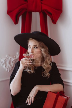 Téléchargez les photos : Belle femme élégante en robe de fête noire et chapeau avec verre de champagne et cadeaux du Nouvel An. Concentration sélective douce. - en image libre de droit