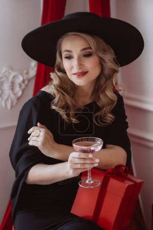 Téléchargez les photos : Belle femme élégante en robe de fête noire et chapeau avec verre de champagne et cadeaux du Nouvel An. Concentration sélective douce. - en image libre de droit