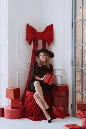 Foto de Hermosa mujer elegante en un vestido de fiesta negro y sombrero entre una gran cantidad de regalos rojos de Año Nuevo. Enfoque selectivo suave. - Imagen libre de derechos