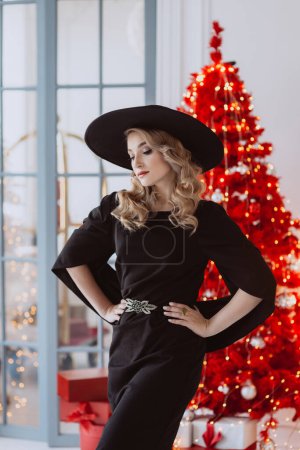 Photo for Beautiful elegant woman in a black festive dress and hat on the background of a red Christmas tree. Soft selective focus. - Royalty Free Image