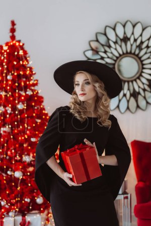 Foto de Hermosa mujer elegante en un vestido negro festivo y sombrero con un regalo de Año Nuevo en el fondo de un árbol de Navidad rojo. Enfoque selectivo suave. - Imagen libre de derechos