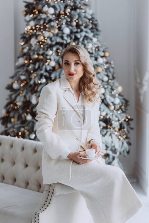 Photo for Young happy european businesswoman with cup of coffee with decorated Christmas tree behind, woman working in home office during winter holidays. Soft selective focus. - Royalty Free Image