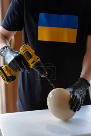 Téléchargez les photos : Le chef en gants noirs ouvre un œuf d'autruche avec une perceuse. Concentration sélective douce. - en image libre de droit