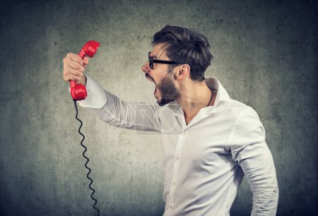 Téléchargez les photos : Vue latérale du jeune homme barbu décontracté tenant un récepteur téléphonique rouge et criant de colère. - en image libre de droit