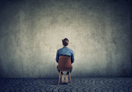 Un jeune homme s'assoit sur une chaise et regarde un mur de béton vide