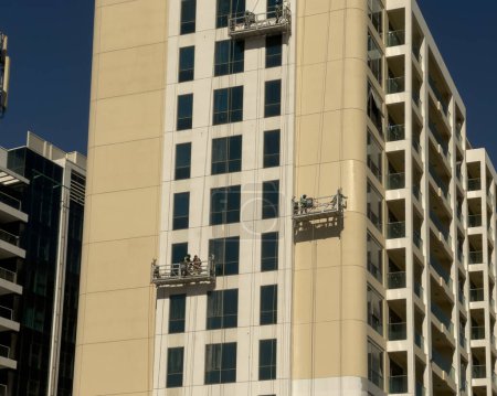 Téléchargez les photos : Dubaï, Émirats arabes unis - 7 janvier 2022 : Des travailleurs de la construction peignent la façade extérieure d'un bâtiment résidentiel, abaissés sur des échafaudages suspendus suspendus sur le côté du bâtiment. - en image libre de droit