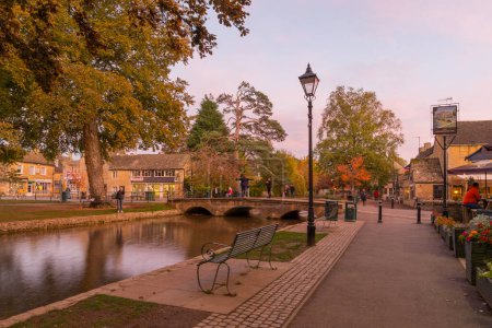 Photo for Bourton-on-the-Water, UK - October 17, 2022: Sunset scene of typical houses, the river Windrush, locals and visitors, in the village Bourton-on-the-Water, the Cotswolds region, England, UK - Royalty Free Image