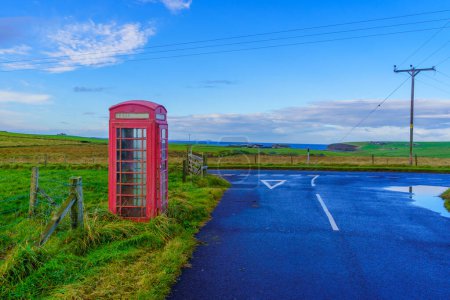 Blick auf eine rote Telefonzelle in einer Landschaft aus Feldern mit Straße auf den Orkney-Inseln, Schottland, Großbritannien