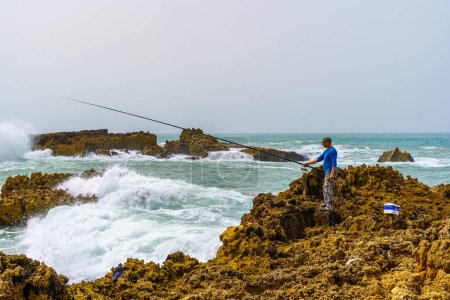 Téléchargez les photos : Oualidia, Maroc - 08 avril 2023 : Vue d'un pêcheur, et vagues s'écrasant sur la plage rocheuse, dans la station balnéaire du village Oualidia, la côte Atlantique du Maroc - en image libre de droit