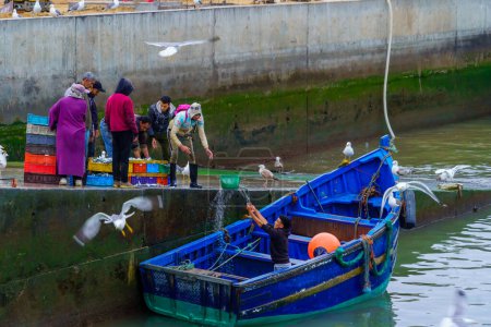 Téléchargez les photos : Essaouira, Maroc - 07 avril 2023 : Scène du lever du soleil du port de pêche, avec des pêcheurs livrant des paniers de sardines, à Essaouira (Mogador), Maroc - en image libre de droit
