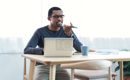 Photo for African American bearded freelancer Man records audio message holds phone looks aside. Black Entrepreneur speaking online technology and virtual assistant note at home. Mobile ai technology concept - Royalty Free Image