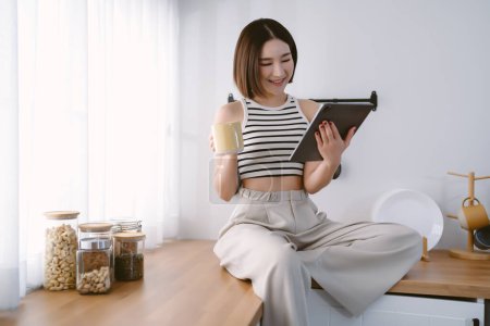Photo for Smiling beautiful Asian woman sits and relaxes on top counter kitchen using tablet browsing unlimited wireless internet in kitchen. Korea girl influencer or blogger streaming on social network online. - Royalty Free Image