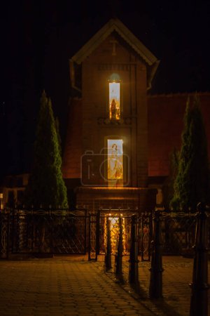 Téléchargez les photos : Chapelle illuminée dans une petite ville, vue de nuit, figurines surlignées de saints - en image libre de droit
