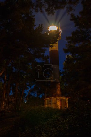 Photo for Lighthouse on the coast among the trees view at night, light beam for navigation - Royalty Free Image