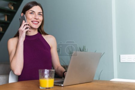 Freiberufliche Frau, die mit dem Handy am Laptop tippt und vom Homeoffice aus arbeitet. Glückliches Mädchen, das im Homeoffice arbeitet. Online-Fernstudium und Arbeit.