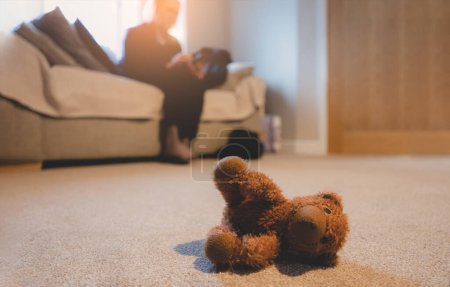 Foto de Osito de peluche acostado en una alfombra con una mujer borrosa sentada en un sofá, oso de peluche solitario acostado solo en la sala de estar, concepto solitario, violencia doméstica, Día Internacional de los Niños Desaparecidos. - Imagen libre de derechos