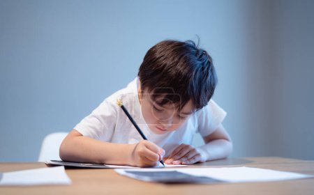 Enfant assis sur la table faisant ses devoirs, enfant garçon tenant un stylo noir écrivant sur du papier blanc, jeune garçon pratiquant des mots anglais à la maison. École primaire et enseignement à domicile, concept d'enseignement à distance 