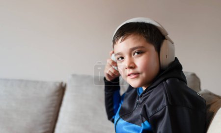 Lächelnder kleiner Junge mit Kopfhörer, der online lernt, Homeschooling-Konzept, Fröhliches Kind mit drahtlosem Headset, das Musik hört und in die Kamera schaut, Home Bildungstechnologie und Musikkonzept