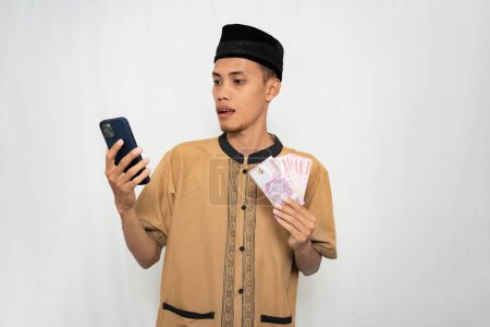 Foto de Hombre musulmán asiático vistiendo ropa musulmana se sorprende mirando la pantalla del teléfono inteligente mientras lleva dinero en su otra mano. Fondo blanco aislado. - Imagen libre de derechos
