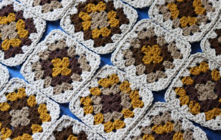 Crocheted granny squares made of natural brown, mustard and beige wool. Soft and fluffy crochet ornament on blue textured background. 