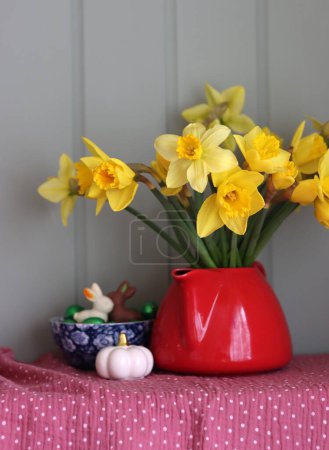 Bouquet of golden daffodils in red teapot on a light grey wooden background with copy space.Still life with red teapot and daffodils on sage green background.