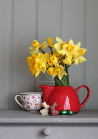 Photo for Bouquet of golden daffodils in red teapot on a light grey wooden background with copy space.Still life with red teapot and daffodils on sage green background. - Royalty Free Image