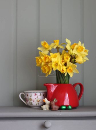 Bouquet of golden daffodils in red teapot on a light grey wooden background with copy space.Still life with red teapot and daffodils on sage green background.