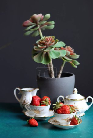 Erdbeeren in Tassen und Vase mit Sukkulente auf grünem Tisch. Dunkler Hintergrund mit Kopierraum. Stillleben mit reifen Sommerbeeren. Ausgewogenes Ernährungskonzept. 