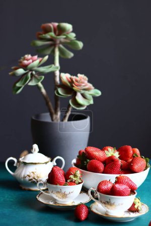 Erdbeeren in Tassen und Vase mit Sukkulente auf grünem Tisch. Dunkler Hintergrund mit Kopierraum. Stillleben mit reifen Sommerbeeren. Ausgewogenes Ernährungskonzept. 