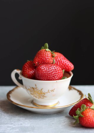 Erdbeeren in einer kleinen weiß-goldenen Porzellantasse auf dunklem Hintergrund mit Platz für Text. Stillleben-Foto mit Retro-Geschirr und Sommerfrüchten. Frisches Konzept.