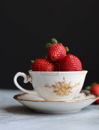 Erdbeeren in einer kleinen weiß-goldenen Porzellantasse auf dunklem Hintergrund mit Platz für Text. Stillleben-Foto mit Retro-Geschirr und Sommerfrüchten. Frisches Konzept.