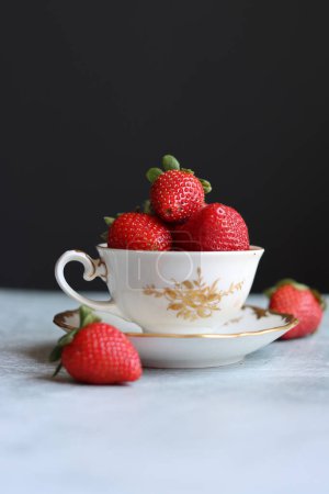 Strawberries in a small white and gold porcelain cup on dark background with space for text. Still life photo with retro tableware and summer fruit. Eating fresh concept.