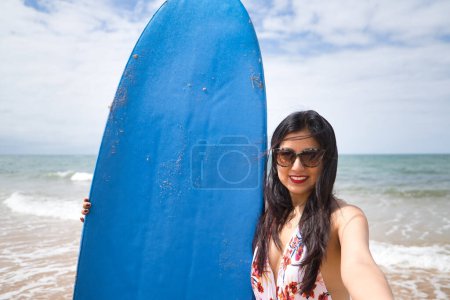 Photo for South American woman, young and beautiful, brunette with sunglasses and swimsuit, taking a selfie while holding a blue surfboard. Concept sea, sand, sun, beach, vacation, surf, summer. - Royalty Free Image